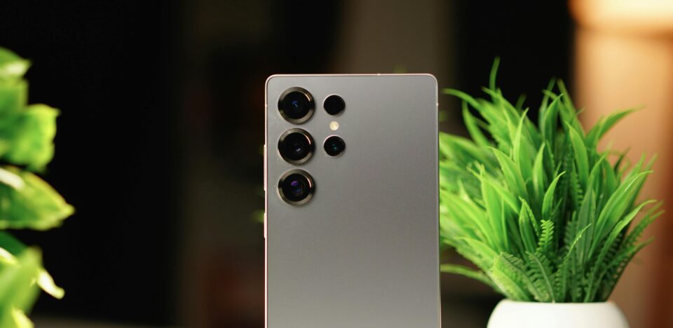 Modern smartphone standing on a desk with green potted plants in a warm indoor setting.