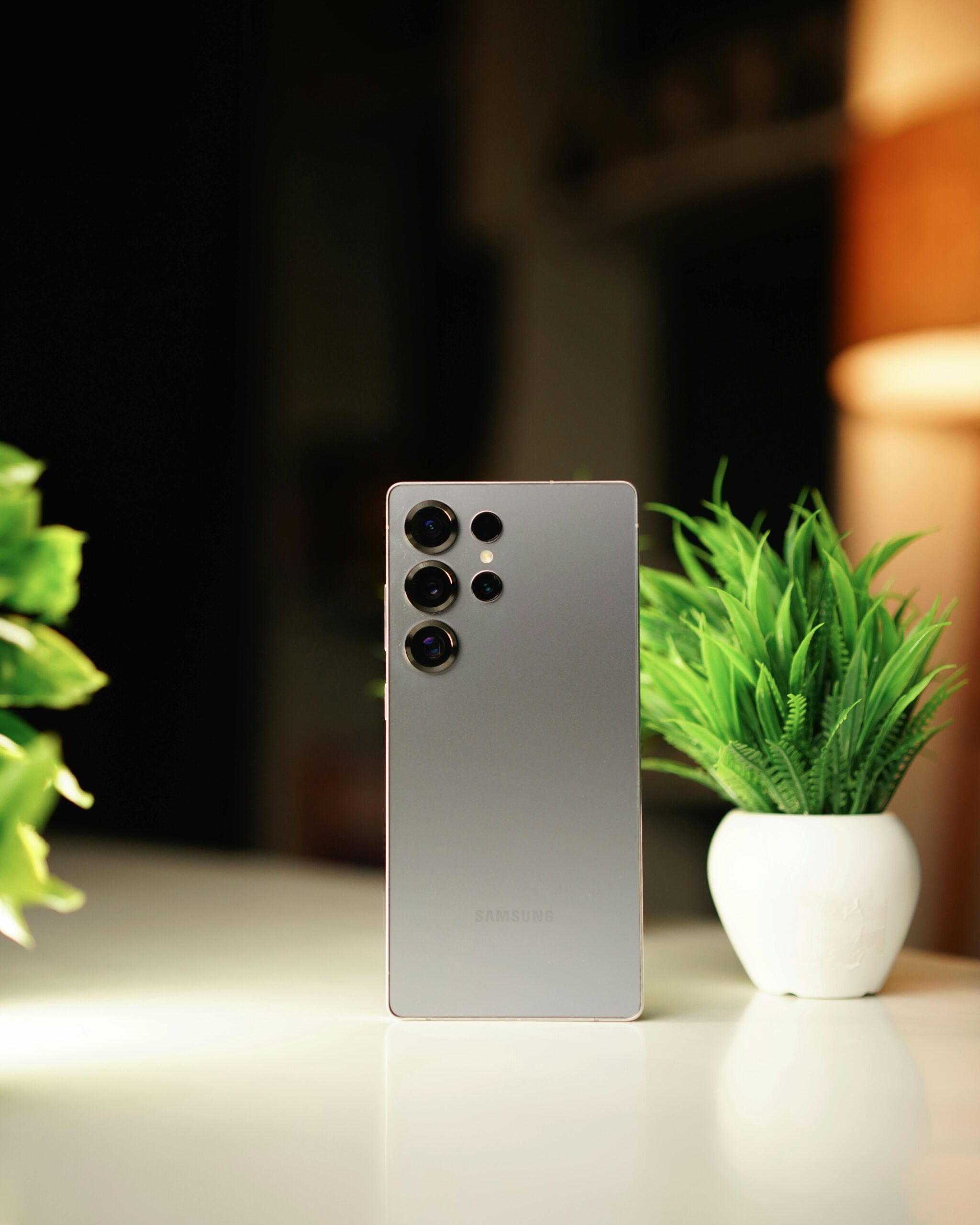 Modern smartphone standing on a desk with green potted plants in a warm indoor setting.